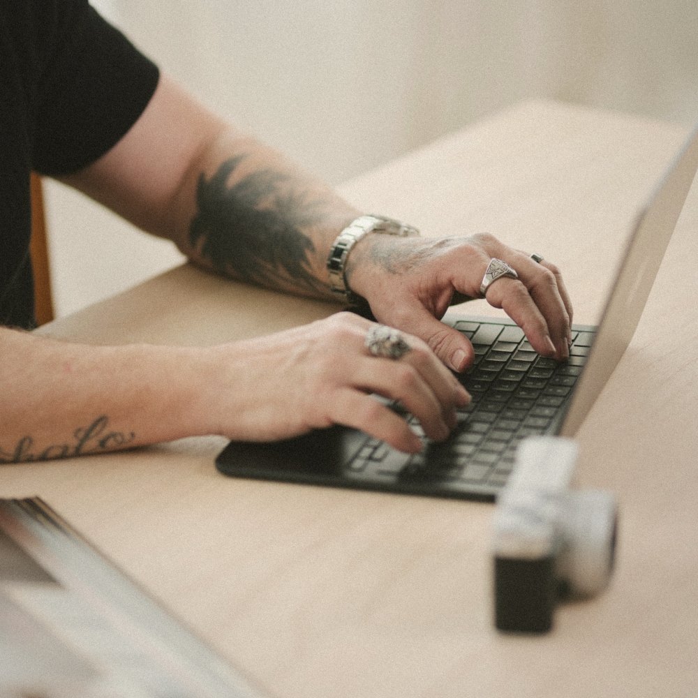 Close-up of John Lyons's tattooed hands typing on a laptop beside a vintage camera, printed as an instant photo.